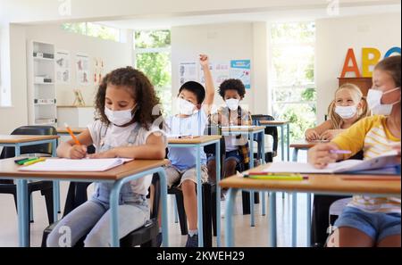 I bambini imparano in classe dopo una pandemia covida, indossando maschere protettive per il viso. Bambini piccoli seduti a scuola con braccio sollevato per chiedere Foto Stock