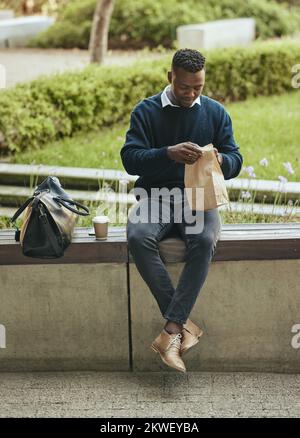 Uomo d'affari che mangia cibo per pranzo durante la sua pausa all'aperto in un parco che apre una borsa di carta marrone. Affamato, buon maschio nero tempo per mangiare e bere fuori Foto Stock