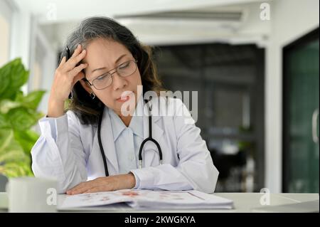 Stressato medico asiatico anziano che soffre di mal di testa o emicrania mentre lavora alla sua scrivania ufficio. Foto Stock