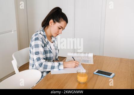 Giovane donna che fa i compiti a casa mentre si siede al tavolo in cucina a casa Foto Stock
