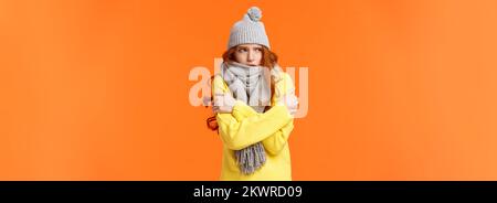 Il suo freddo freddo all'esterno. Cute ragazza redhead scuotendo e abbracciandosi per riscaldarsi, aspettando qualcuno sulla strada in sciarpa grigia, cappello invernale durante la neve Foto Stock