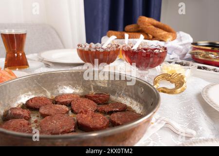 Foto di primo piano con messa a fuoco selettiva della salsiccia fritta chiamata sucuk in padella. Tradizionale tavolo per la colazione turco. Bicchiere di tè, vasetto, bagel chiamati simit. Foto Stock