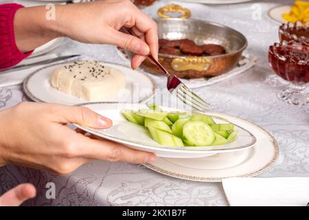 Donna che tiene e serve piatto di cetriolo a fette. Vista ravvicinata. Salsiccia fritta chiamata sucuk sullo sfondo. Formaggio bianco e marmellata sul tavolo. Foto Stock
