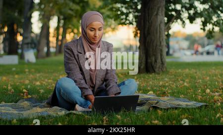 Musulmano ragazza studentessa islamica in hijab business donna freelancer utente utilizza laptop seduto su erba verde nel parco di lavoro a distanza studiare in estate Foto Stock