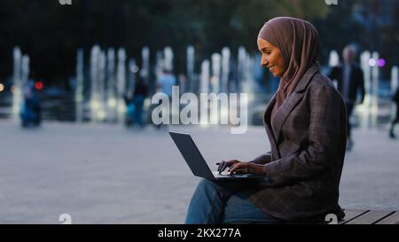 Occupato successo musulmano studente ragazza in hijab islamico business donna freelancer utilizzando laptop app lavorando in città fontane sfondo digitando e-mail in Foto Stock