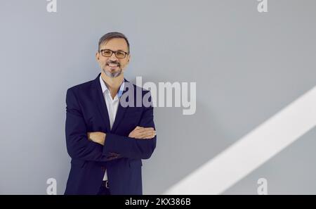 Ritratto di successo uomo d'affari maturo in tuta e occhiali in piedi vicino alla parete in ufficio. Foto Stock