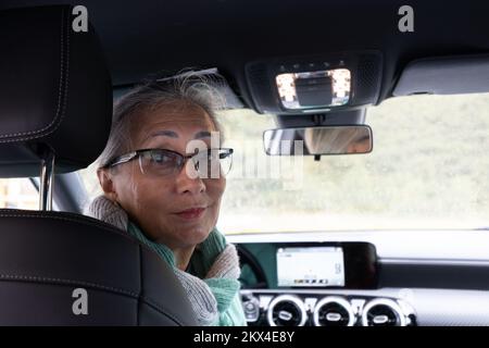 Una signora dai capelli grigi siede sul sedile del conducente di un'auto e guarda intorno ai passeggeri. Foto Stock