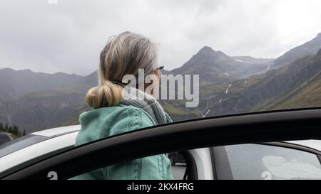 Una donna dai capelli grigi in un caldo maglione si trova vicino ad un'auto in una valle alpina e guarda le montagne. Foto Stock