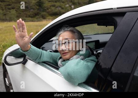 Una signora dai capelli grigi guarda fuori dal finestrino di un'auto e le fa ondare la mano Foto Stock