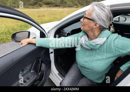 Una signora dai capelli grigi siede sul sedile del conducente in un'auto con una porta aperta e guarda lontano Foto Stock