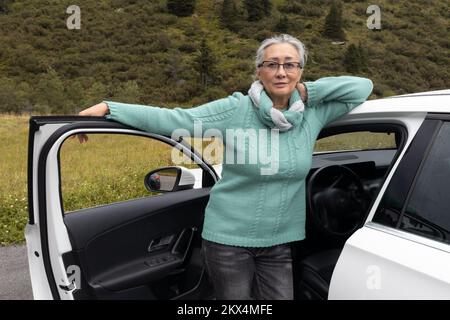 Una donna dai capelli grigi in un maglione caldo si alza alla porta dell'auto aperta e guarda la telecamera Foto Stock