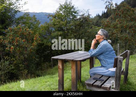 Una donna dai capelli grigi siede su un tavolo di legno, in un ambiente alpino. Foto Stock