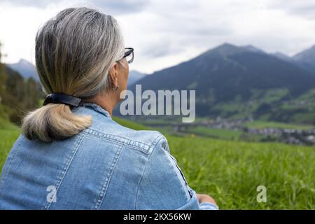 Una donna anziana in denim si siede su un alto prato e guarda le montagne sul lato opposto della gola. Foto Stock