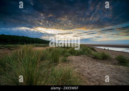 Una coppia cammina lungo la spiaggia e le dune di sabbia durante il tramonto a Holkham Beach, Norfolk, Inghilterra, Regno Unito. Foto Stock