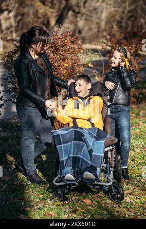 Diversità e inclusione. Felice famiglia, madre, figlia e figlio ragazzo con paralisi cerebrale trascorrere insieme il tempo in autunno autunno parco autunno. Ragazzo teen che Foto Stock