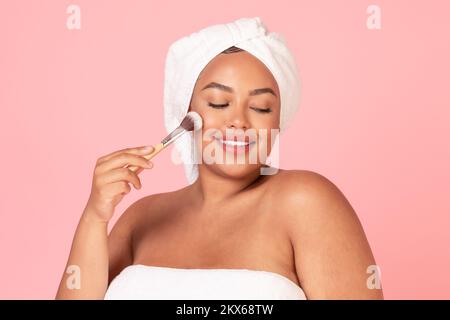 Attraente giovane donna chubby con spazzola per il trucco in posa avvolto in asciugamano, godendo di nuovi prodotti per il trucco Foto Stock