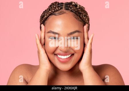 Ritratto di felice bella donna afroamericana che tocca il suo volto e sorridendo alla macchina fotografica, posando su sfondo rosa Foto Stock