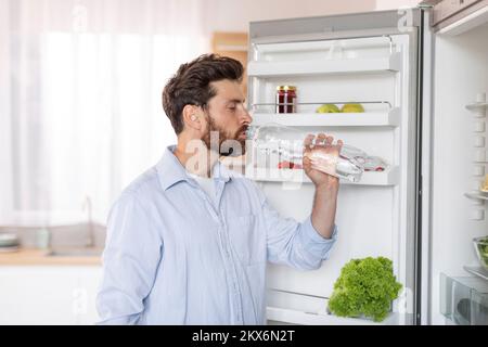 Sete adulta uomo caucasico con barba in camicia bianca apre la porta del frigorifero, beve acqua dalla bottiglia Foto Stock