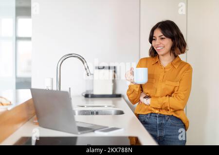 Giovane donna araba sorridente che usa il laptop e bere caffè in cucina Foto Stock