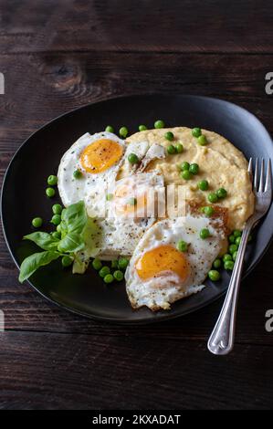 Polenta cremosa con uova fritte, lato soleggiato e piselli verdi su piatto scuro su tavolo di legno Foto Stock
