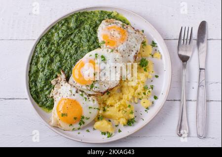 Spinaci cremati con uova fritte e patate su un piatto isolato su fondo bianco. Giacitura piatta Foto Stock