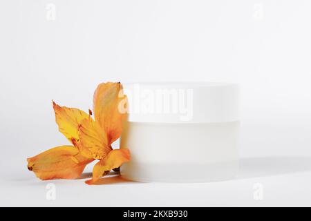 Vasetto cosmetico bianco con crema su fondo bianco con fiore. Cosmetici per la stagione primaverile. Pubblicità di cosmetici primaverili. Cosmetologia o cura della pelle Foto Stock