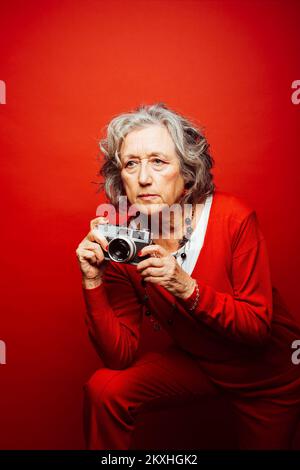 Donna anziana che indossa abiti rossi, fotografa con una vecchia macchina fotografica, su sfondo rosso Foto Stock