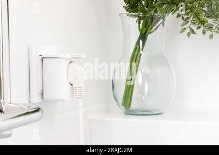 Presa elettrica europea bianca con presa intelligente inserita in un bagno moderno e luminoso con parete piastrellata bianca e bouquet di plastica verde in vetro v Foto Stock