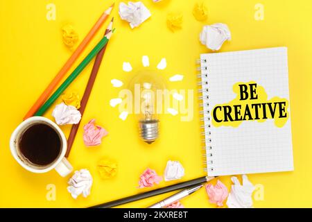 Concetto di business e di istruzione. Su uno sfondo giallo, una tazza di caffè, una lampadina, matite, un notebook con l'iscrizione - SIATE CREATIVI Foto Stock