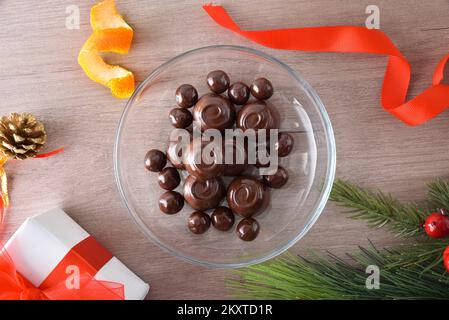 Caramelle al cioccolato in un piatto rotondo di vetro trasparente al centro di un tavolo di legno con decorazioni natalizie intorno. Vista dall'alto. Foto Stock