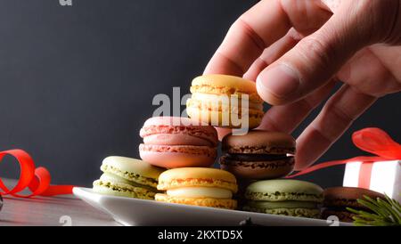 Raccogliere a mano un macaron al cioccolato da un piatto pieno di sapori diversi su un tavolo di legno e sfondo scuro. Vista frontale. Foto Stock