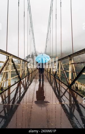 Ragazza con ombrello blu su ponte sospeso in ferro bagnato in caso di pioggia, vista posteriore. Foto Stock