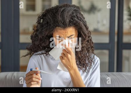 La donna afro-americana è molto malata misura la temperatura, si sente molto male. Una giovane femmina ammalata soffia il naso in un tovagliolo Foto Stock