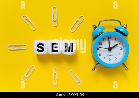 Finanza ed economia. Su sfondo giallo, un orologio di allarme blu, graffette e cubi bianchi su cui è scritto il testo - SEM Foto Stock