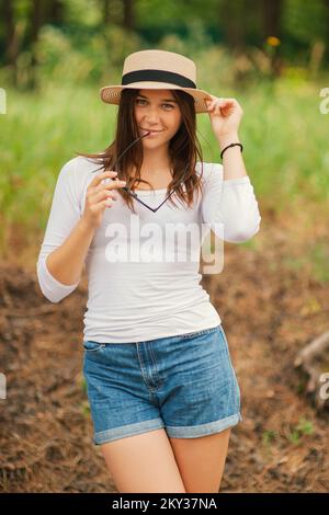 Carino giovane donna alla moda indossando un cappello di regolazione di estate tenendo gli occhiali da sole nelle sue mani a guardare la macchina fotografica in piedi all'aperto Foto Stock