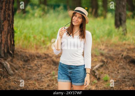 Carino giovane donna alla moda indossando un cappello di regolazione di estate tenendo gli occhiali da sole nelle sue mani a guardare la macchina fotografica in piedi all'aperto Foto Stock