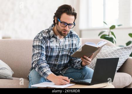 Studente maschile milenne europeo impegnato in abiti domestici con stoppia in occhiali con libro e pc Foto Stock