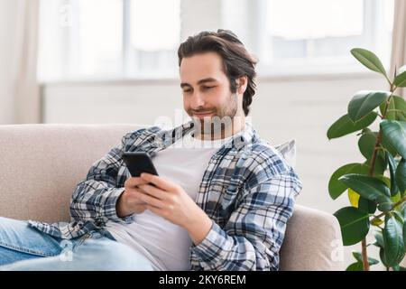 Felice maschio caucasico millenario in abiti domestici con stoppia si trova sul divano e chiacchierando sullo smartphone Foto Stock