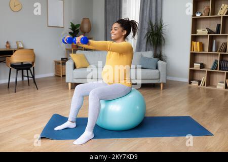 Felice giovane donna incinta nera con pancia grande fare esercizi sulla palla fitness con manubri Foto Stock