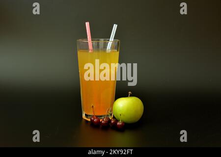 Succo fresco fruttato in un bicchiere alto con cannucce su fondo nero, accanto ad una mela verde matura e ad una manciata di ciliegie. Primo piano. Foto Stock