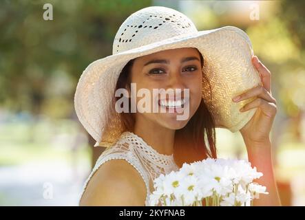La primavera è qui. Una giovane donna che tiene fiori e indossa un cappello da sole in piedi in un parco. Foto Stock