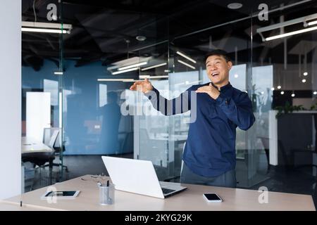 Uomo d'affari che festeggia il successo, uomo asiatico che balla e canta sul posto di lavoro, uomo in camicia che lavora in un ufficio moderno utilizzando un computer portatile al lavoro. Foto Stock
