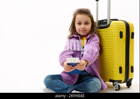 Bella bambina in camicia viola e tute in denim, con biglietto aereo seduto vicino al giallo valise su sfondo bianco Foto Stock