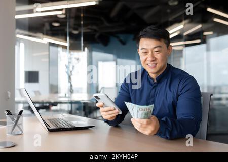 L'uomo d'affari asiatico ha vinto il dollaro, l'uomo seduto all'interno dell'ufficio che tiene contanti e telefono cellulare. Foto Stock