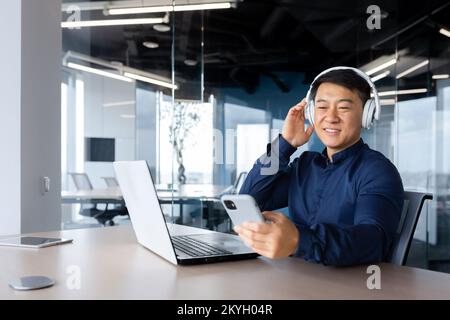 Uomo d'affari che ascolta musica e podcast sul posto di lavoro, uomo asiatico sorridente e felice utilizzando il computer portatile sul lavoro, uomo con cuffie e telefono che ascolta la radio online all'interno dell'ufficio. Foto Stock