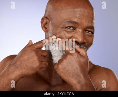 Faccia, bellezza e cura della pelle con un uomo nero anziano che controlla la sua pelle per un pimple o zit in studio su uno sfondo viola. Ritratto, benessere e. Foto Stock