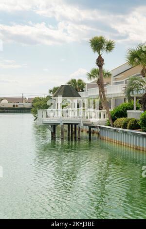 Vista di un gazebo su un ponte sull'acqua salmastra di un lago con parete di contenimento in acciaio a Destin, Florida. Vista sul ponte davanti alle case con balconi. Foto Stock