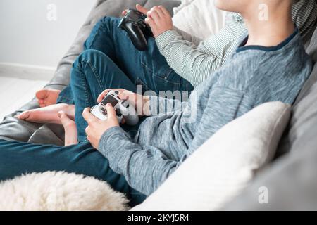 Mani di ragazzi seduti sul divano con joystick di gioco, vista laterale. I bambini giocano a videogiochi. Ragazzi in denim e casual longsleeves relax a casa Foto Stock