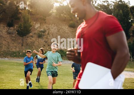 Bambini che corrono in un campo scolastico durante la PE. Gruppo di ragazzi della scuola elementare che hanno una sessione di educazione fisica con il loro allenatore. Sport di divertimento traini Foto Stock
