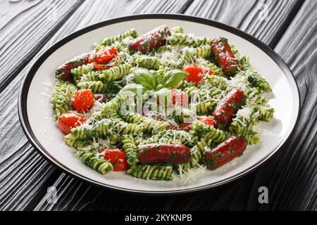 Pranzo abbondante Fusilli pasta con salsicce, pomodori, salsa di pesto verde e formaggio primo piano in un piatto sul tavolo. Orizzontale Foto Stock
